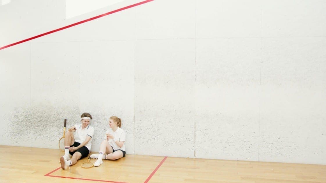 Two squash players resting against the back wall of a court